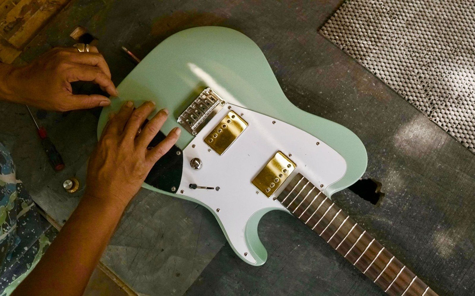 A luthier's workbench with a Fender-style bass neck clamped in a work cradle, truss-rod tools and radius gauges alongside, workshop light from above
